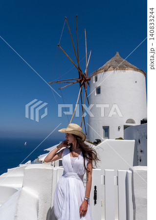 Beautiful young woman in white dress and straw hat posing near traditional windmill in Santorini Greece Beautiful young woman in white dress and straw hat posing near traditional windmill in Santorini Greece 140833244