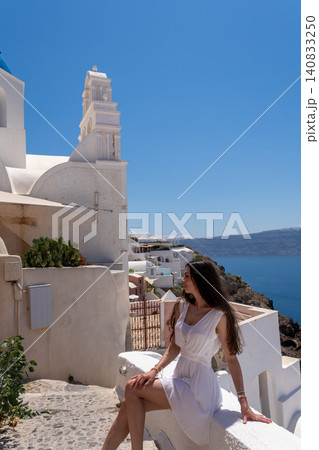 Beautiful young woman in white dress sitting near traditional church bell tower in Santorini Greece Beautiful young woman in white dress sitting near traditional church bell tower in Santorini Greece 140833250