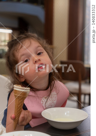 Vertical shot of cute little girl with dirty face eating ice cream in restaurant, funny toddler holding waffle cone at table, messy kid with milk mustache after dessert Vertical shot of cute little girl with dirty face eating ice cream in restaurant, funny toddler holding waffle cone at table, messy kid with milk mustache after dessert 140833312