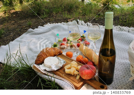 Picnic setup featuring assorted cheeses, croissants, fruits, and white wine on a blanket by a serene lakeside surrounded by greenery and natural light Picnic setup featuring assorted cheeses, croissants, fruits, and white wine on a blanket by a serene lakeside surrounded by greenery and natural light 140833769