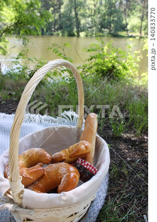 A picnic basket containing various bread rolls and a jar of jam rests on a blanket by a tranquil lake, with trees and grass in the background A picnic basket containing various bread rolls and a jar of jam rests on a blanket by a tranquil lake, with trees and grass in the background 140833770