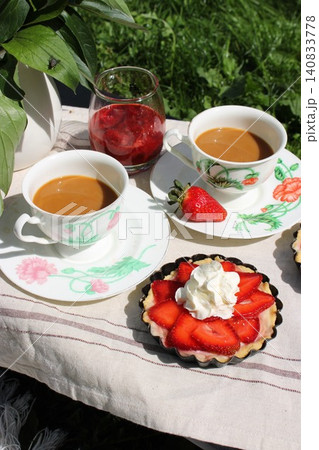 Two cups of coffee with decorative saucers, fresh strawberries, and a strawberry tart on a tablecloth in a garden setting with greenery in the background 140833778