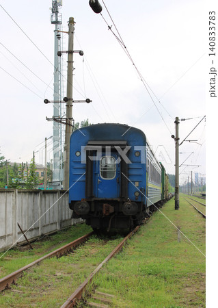 Blue passenger train car parked on railway tracks with green grass and concrete wall in the background, power lines and communication tower visible nearby 140833783