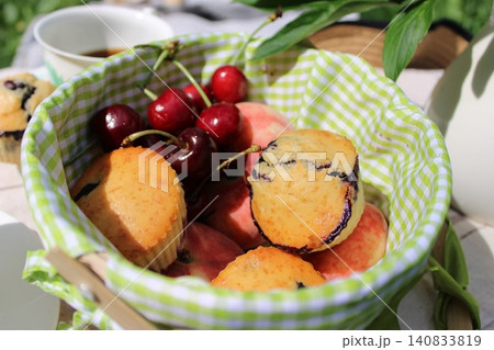 Blueberry muffins and cherries are arranged in a green checkered basket with ripe peaches on a sunny outdoor table, creating a vibrant summer scene 140833819