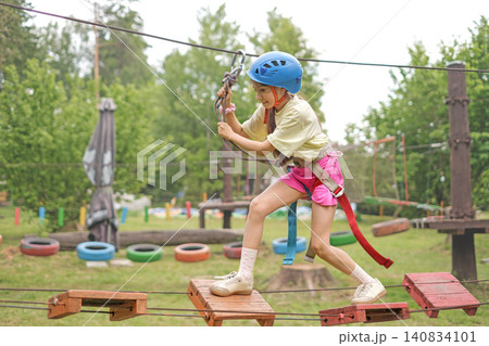 A girl wearing protective sports gear and a helmet is walking along a suspended bridge. Outdoor recreation and entertainment park A girl wearing protective sports gear and a helmet is walking along a suspended bridge. Outdoor recreation and entertainment park 140834101