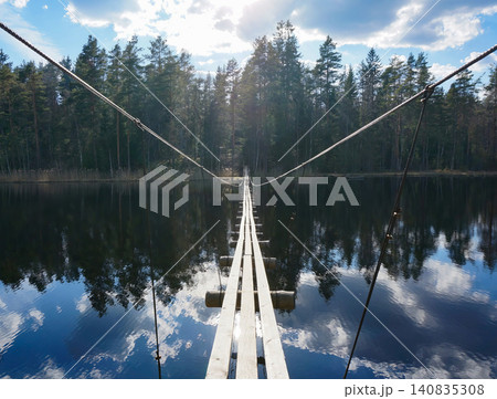 A narrow wooden suspension bridge crossing a calm lake in a forest A narrow wooden suspension bridge crossing a calm lake in a forest 140835308