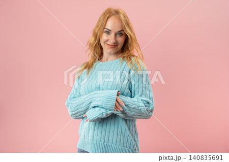 Young woman with red hair wearing a blue knitted sweater, standing confidently against a pink background, showcasing a shy yet warm expression 140835691