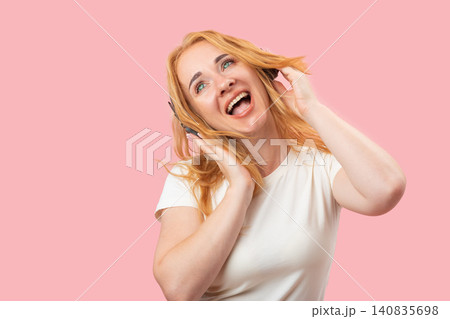 Smiling young woman with blonde hair listening to music through black headphones, expressing joy and energy while looking upwards against a pink background 140835698