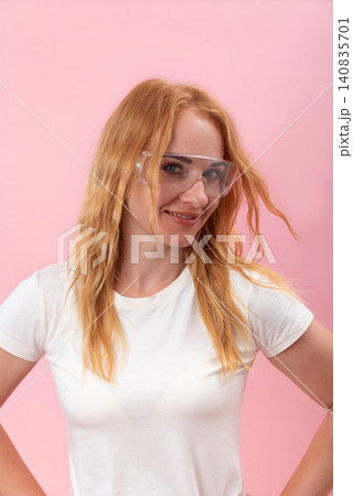 Young redhead woman wearing clear protective eyewear and a white t shirt, standing confidently with a smile against a solid pink background Young redhead woman wearing clear protective eyewear and a white t shirt, standing confidently with a smile against a solid pink background 140835701
