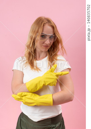 Woman wearing yellow rubber gloves and protective glasses looking at the camera while confidently pointing her gloved hand to the right against a pink background Woman wearing yellow rubber gloves and protective glasses looking at the camera while confidently pointing her gloved hand to the right against a pink background 140835702