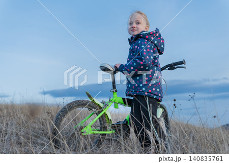 Young blonde girl in a polka dot jacket stands with her bicycle in dry grass, full length, looking away under a wide blue sky, outdoor childhood leisure and freedom Young blonde girl in a polka dot jacket stands with her bicycle in dry grass, full length, looking away under a wide blue sky, outdoor childhood leisure and freedom 140835761