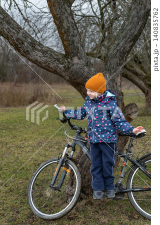 Young girl with orange hat and blue jacket happily standing on a bicycle seat, leaning against a tree trunk in a grassy outdoor setting, exploring and enjoying childhood Young girl with orange hat and blue jacket happily standing on a bicycle seat, leaning against a tree trunk in a grassy outdoor setting, exploring and enjoying childhood 140835762