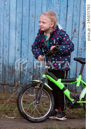 Young girl with braided hair holding her green bicycle and looking away from the camera, reflecting childhood curiosity and outdoor fun against a blue wooden background 140835763