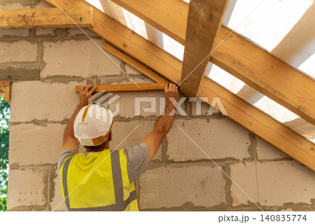 Construction worker wearing a hard hat and safety vest, installing wooden beams for a roof frame on a new residential house building site, focusing on structural elements Construction worker wearing a hard hat and safety vest, installing wooden beams for a roof frame on a new residential house building site, focusing on structural elements 140835774