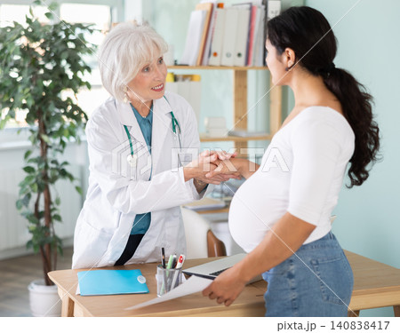 Latin pregnant woman at doctors consultation, patient shakes hands with senior female doctor at table Latin pregnant woman at doctors consultation, patient shakes hands with senior female doctor at table 140838417