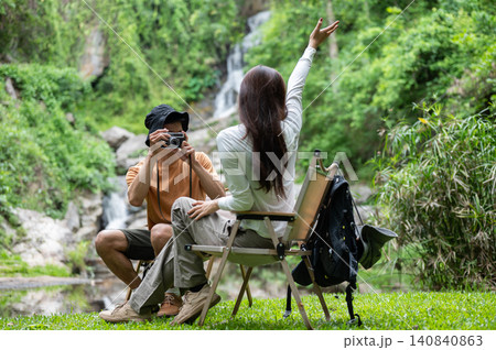 Asian man tourist holding camera taking photo of girlfriend sitting on chair at waterfall in forest. 140840863