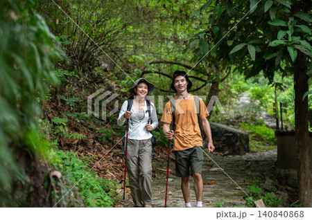 Happy asian male and female hikers walking with staff through lush green forest on a mountain trail Happy asian male and female hikers walking with staff through lush green forest on a mountain trail 140840868