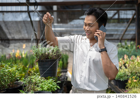 Asian man visitor holding a hanging plant while talking on phone in greenhouse nursery garden center Asian man visitor holding a hanging plant while talking on phone in greenhouse nursery garden center 140841526