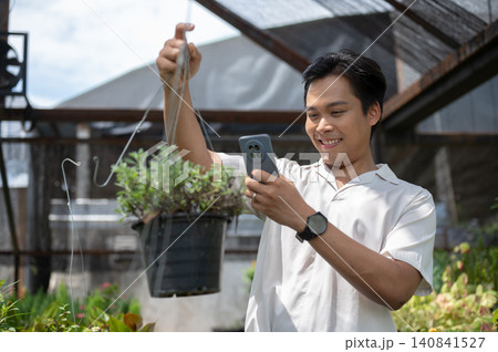 Asian man visitor holding a hanging plant and talking on phone in a greenhouse nursery garden center Asian man visitor holding a hanging plant and talking on phone in a greenhouse nursery garden center 140841527