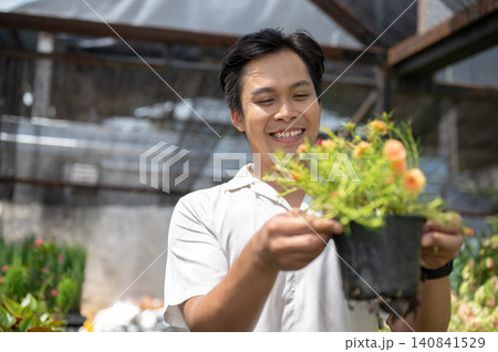 Close up of smiling asian man holding a flowering potted plant in a greenhouse nursery garden center Close up of smiling asian man holding a flowering potted plant in a greenhouse nursery garden center 140841529