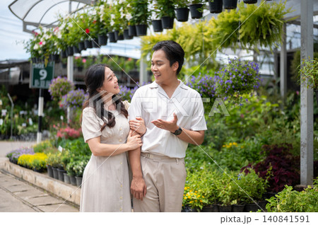 Asian man and woman couple linking arms chatting together beside colorful plants in nursery garden. Asian man and woman couple linking arms chatting together beside colorful plants in nursery garden. 140841591