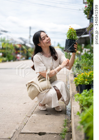Pretty asian woman examining potted plant while sitting next to flower bed at nursery garden center. Pretty asian woman examining potted plant while sitting next to flower bed at nursery garden center. 140841599