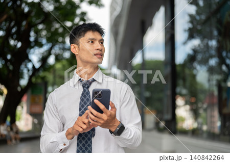 Asian businessman office worker holding smartphone and looking away thoughtfully outside a building Asian businessman office worker holding smartphone and looking away thoughtfully outside a building 140842264
