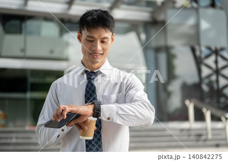 Asian businessman office worker checking time on watch while holding coffee cup outside the building Asian businessman office worker checking time on watch while holding coffee cup outside the building 140842275