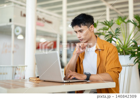 Asian man focused on browsing online with laptop while sitting at table in a contemporary cafe. Asian man focused on browsing online with laptop while sitting at table in a contemporary cafe. 140842309