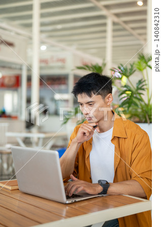 Asian man focused on surfing the internet with laptop while sitting at table in a contemporary cafe. Asian man focused on surfing the internet with laptop while sitting at table in a contemporary cafe. 140842310