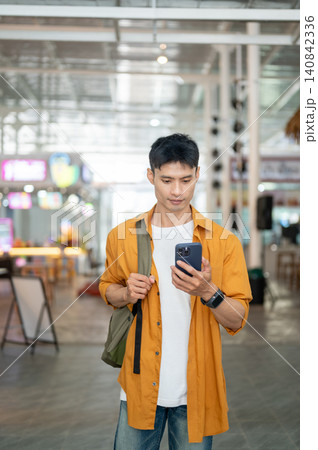Asian man tourist checking smartphone while standing or walking in shopping center with shoulder bag Asian man tourist checking smartphone while standing or walking in shopping center with shoulder bag 140842336