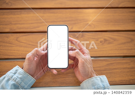 Close up of a man hands holding blank white screen smartphone over wooden table, ideal app template. Close up of a man hands holding blank white screen smartphone over wooden table, ideal app template. 140842359