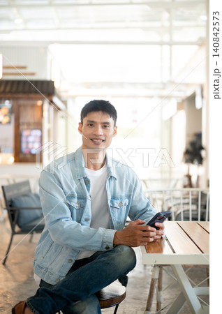 Asian man customer in denim and jeans sitting at table in cafe with smartphone showcasing lifestyle. Asian man customer in denim and jeans sitting at table in cafe with smartphone showcasing lifestyle. 140842573