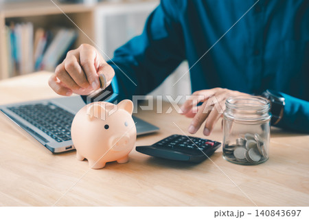 Budget planner savings with piggybank finance bank financial planning, man puts coin in bank while using calculator and laptop at desk for future money investment strategy and goals Budget planner savings with piggybank finance bank financial planning, man puts coin in bank while using calculator and laptop at desk for future money investment strategy and goals 140843697