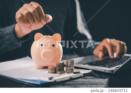 Responsible person putting money in piggy bank for retirement, financial future assurance and wealth, managing economic saving plus loan and finance planning while using laptop at desk Responsible person putting money in piggy bank for retirement, financial future assurance and wealth, managing economic saving plus loan and finance planning while using laptop at desk 140843711