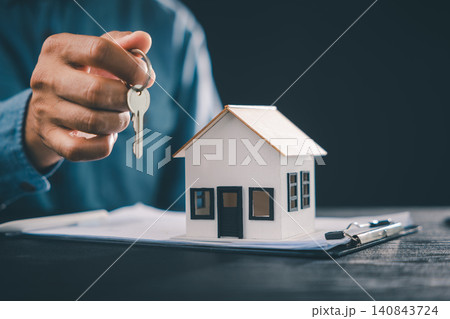 Real estate home purchase involves hand holding keys next to house model, representing mortgage contract, property ownership, residential investment and housing finance agreement Real estate home purchase involves hand holding keys next to house model, representing mortgage contract, property ownership, residential investment and housing finance agreement 140843724