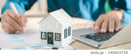 Real estate contract property mortgage finance signing approved, buyer reviews home buying document by house model on desk while using laptop computer for homeowner insurance investment plan 140843739