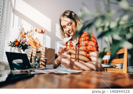 Picture of serious concentrated young Caucasian female artist sitting at desk with painting accessories, holding tube of oil paint, mixing colors on palette; unfinished painting on canvas near her Picture of serious concentrated young Caucasian female artist sitting at desk with painting accessories, holding tube of oil paint, mixing colors on palette; unfinished painting on canvas near her 140844328