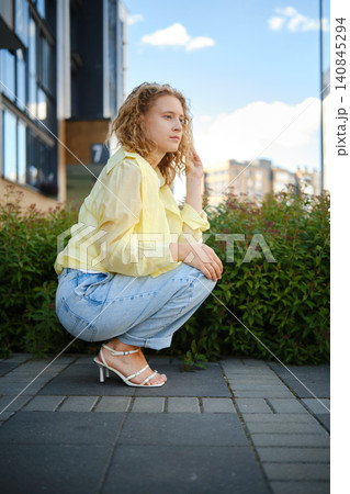 Woman in shirt and jeans crouching in city street during daytime with buildings and plants around Woman in shirt and jeans crouching in city street during daytime with buildings and plants around 140845294