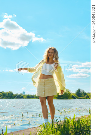 Woman stands by lake wearing yellow shirt and skirt on sunny day with clouds in sky Woman stands by lake wearing yellow shirt and skirt on sunny day with clouds in sky 140845321