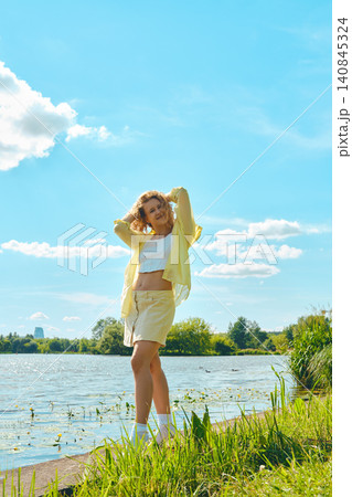 Young woman enjoying sunny day by lake in park with green grass and clouds in blue sky 140845324