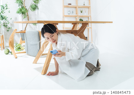 テーブルの下に潜ってスマホを見る女性。地震、緊急地震速報、アラート、防災、災害、怖い、怯える 140845473