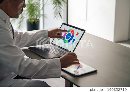 ノートパソコンとタブレットPCを使って作業する白衣の男性。医師、科学、研究、開発、DX、IT ノートパソコンとタブレットPCを使って作業する白衣の男性。医師、科学、研究、開発、DX、IT 140852719