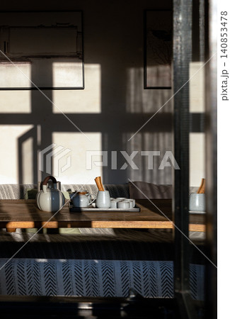 a traditional tea set bathed in sharp, geometric slivers of afternoon sun. The clean lines of the wooden furniture, paired with understated framed artwork and a monochrome patterned bench, evoke a 140853478