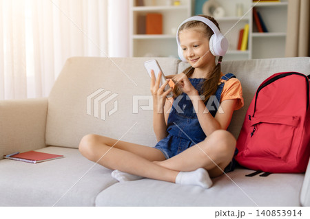 Teen girl wearing headphones using smartphone while sitting on sofa at home Teen girl wearing headphones using smartphone while sitting on sofa at home 140853914