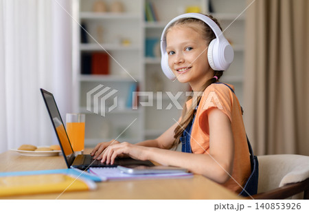 Teen girl wearing headphones working on laptop at home, looking at camera 140853926