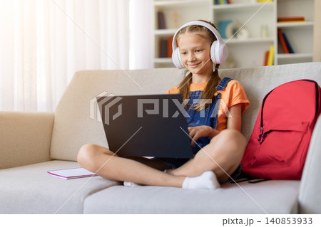 Teen girl in headphones smiling while using laptop on sofa at home Teen girl in headphones smiling while using laptop on sofa at home 140853933