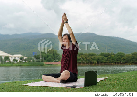 Asian senior man practicing yoga outdoors with laptop online class Asian senior man practicing yoga outdoors with laptop online class 140854301