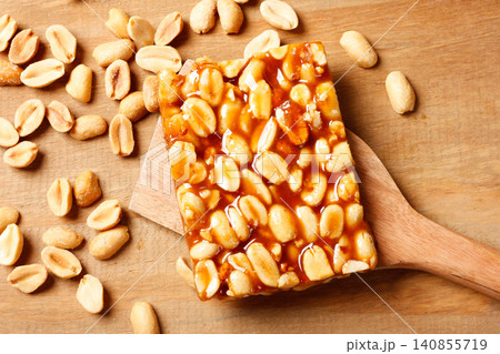 Homemade Brazilian pe de moleque with caramelized peanuts, presented on a wooden spatula and surrounded by roasted peanuts on a rustic surface Homemade Brazilian pe de moleque with caramelized peanuts, presented on a wooden spatula and surrounded by roasted peanuts on a rustic surface 140855719