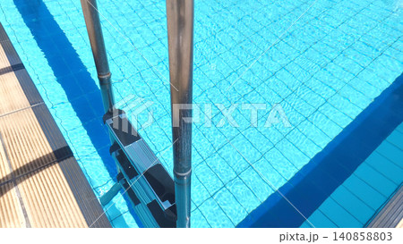 view of a stainless steel staircase leading into the clear blue water of a pool with tiled floors and walls, embodying the concept of swimming, summer relaxation, and recreation view of a stainless steel staircase leading into the clear blue water of a pool with tiled floors and walls, embodying the concept of swimming, summer relaxation, and recreation 140858803